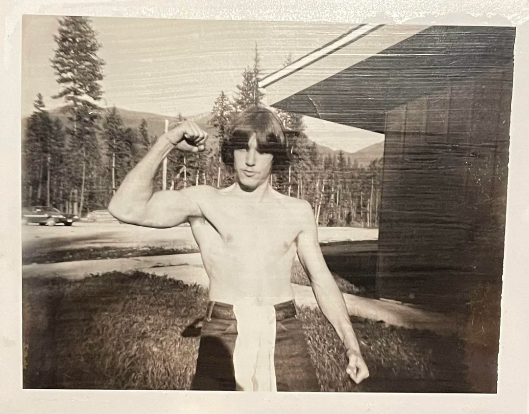 Black-and-white photo of a shirtless young man flexing his right arm, standing outside near a building with trees and mountains in the background. He is wearing dark pants with a light-colored belt or sash.