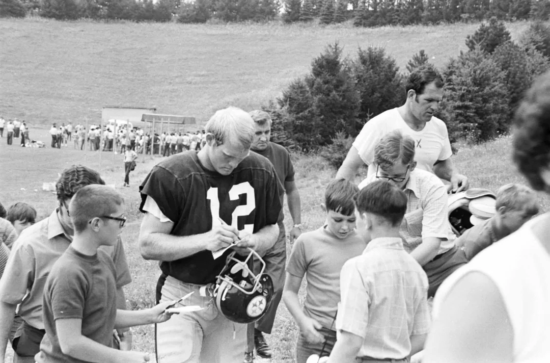 A football player wearing a number 12 jersey signs autographs for a group of children outdoors, with trees and a field in the background. Several other adults and kids are also gathered around him.