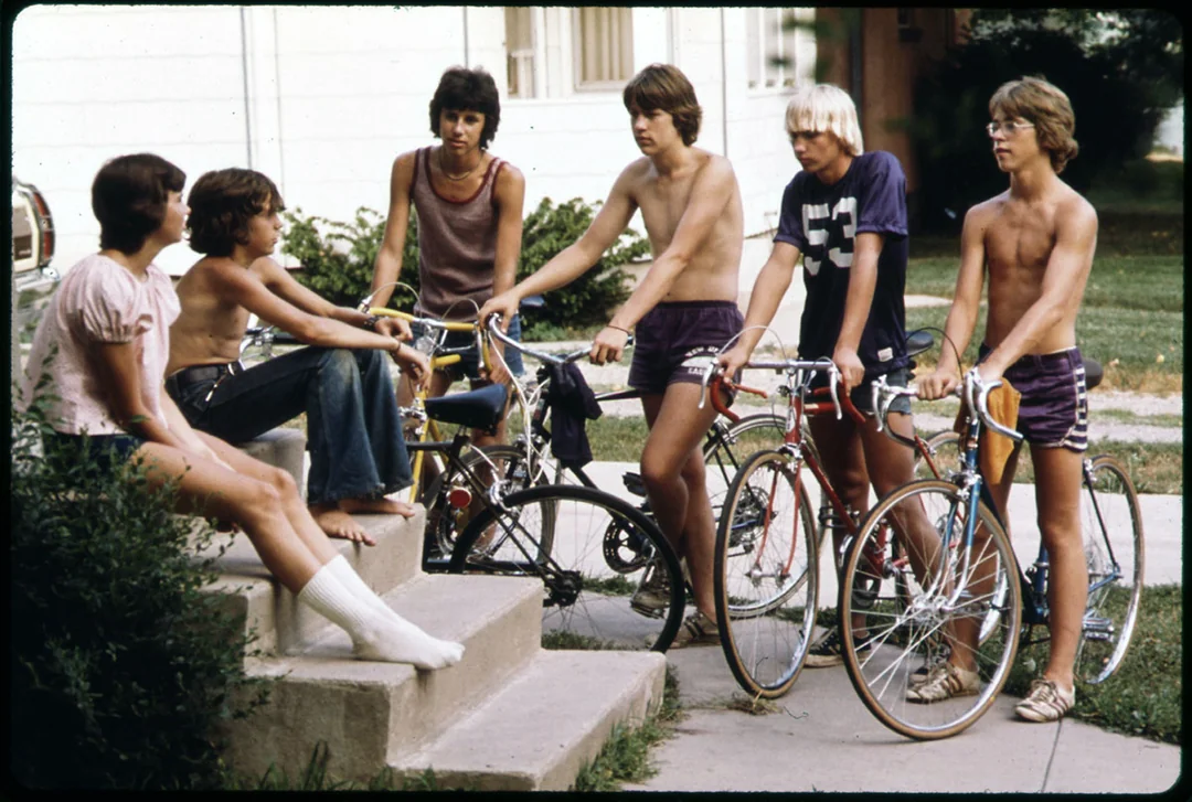 A group of six teenage boys, some shirtless, gather outside on a sunny day with their bicycles. Two sit on concrete steps, while four stand or lean with their bikes on a sidewalk, chatting casually.