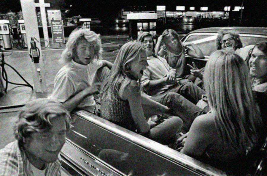 A group of young people sit and laugh together in the back of a pickup truck at a gas station at night. The scene is in black and white, capturing a candid and lively moment among friends.