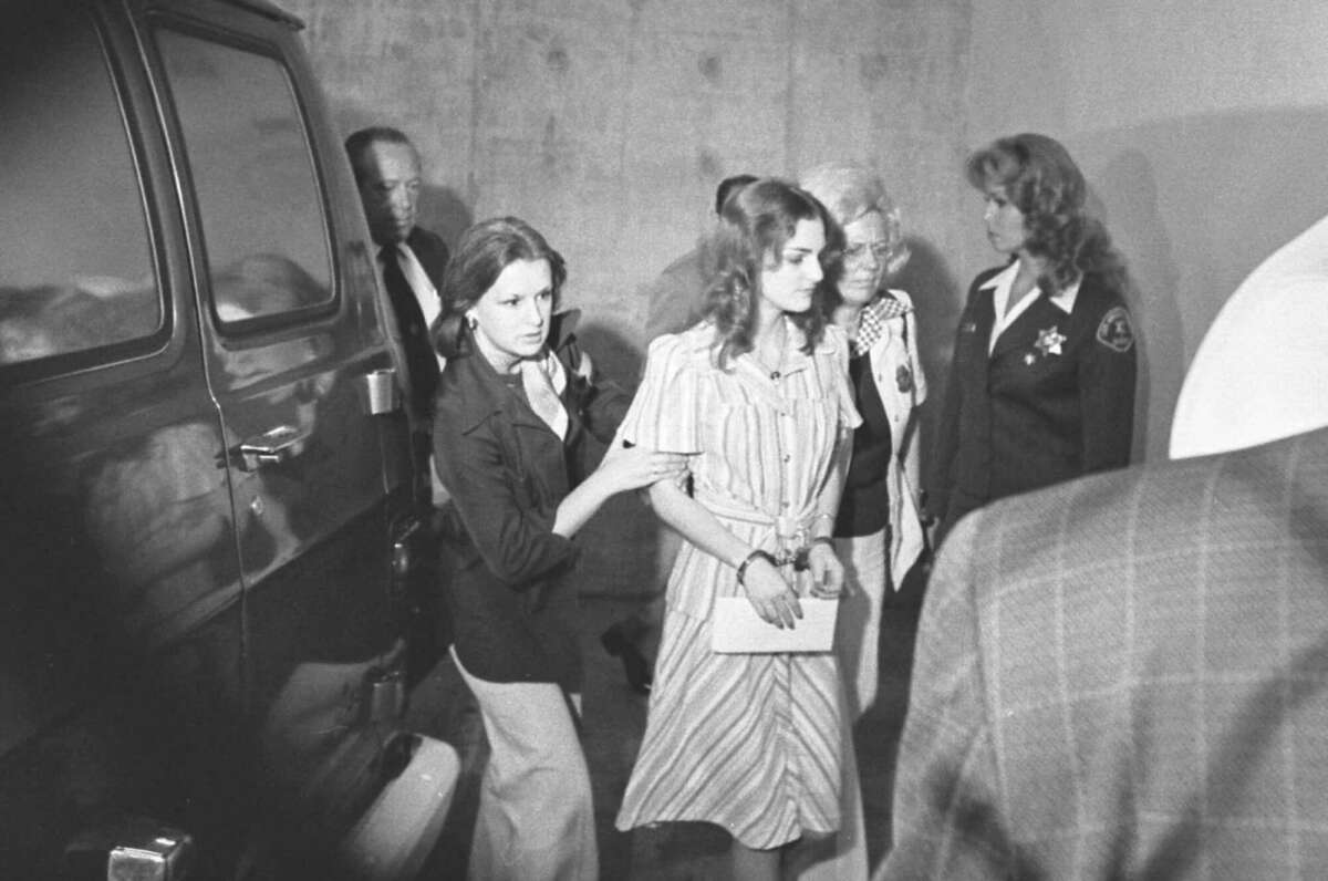 A young woman in a striped dress is escorted by officials, including a uniformed officer, near a dark vehicle as others watch in a dimly lit area.