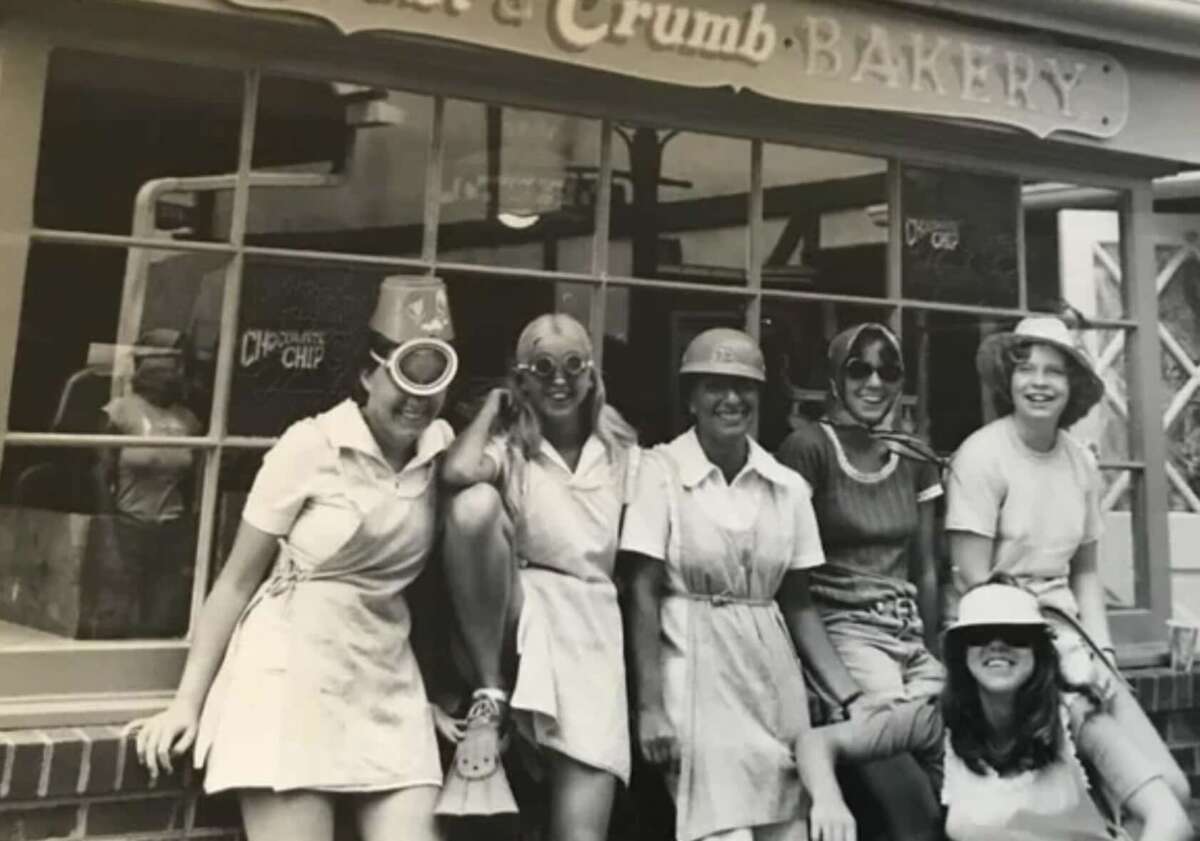 Six women wearing vintage pilot-themed costumes and hats pose and smile in front of a bakery window. The bakery sign above them reads “Crumb Bakery.” It's a black and white photo with a fun, nostalgic atmosphere.