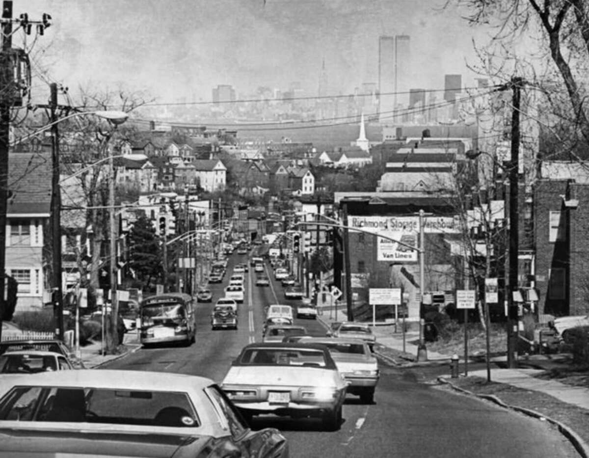 A black-and-white photo of a busy street lined with cars and houses, leading toward the distant New York City skyline, where the Twin Towers are clearly visible against a hazy sky.