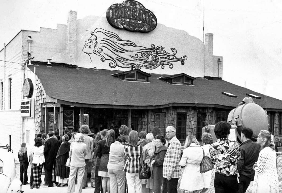 A black-and-white photo shows a long line of people waiting outside a shingled building with a sign reading “Pizza & Pipes” and an artistic mural of a woman with flowing hair on the roof.