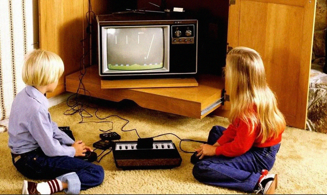 Two children sit on a carpeted floor playing a retro video game console connected to an old television, with a black-and-white game showing on the screen.