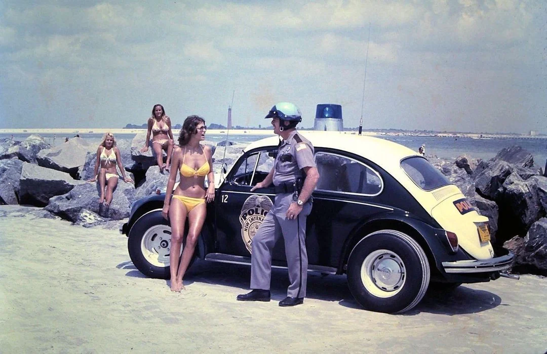 A police officer stands by a vintage police car on a rocky beach, talking to a woman in a yellow bikini while three other women in bikinis walk nearby under a partly cloudy sky.
