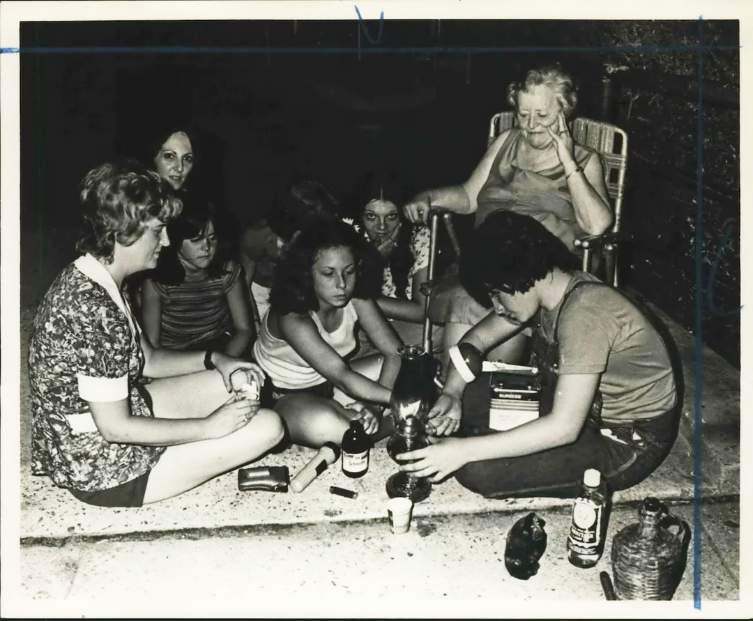 A group of young people and an older woman sit in a circle on the ground, some pouring liquid from a bottle, surrounded by bottles and containers, in a casual outdoor setting at night.