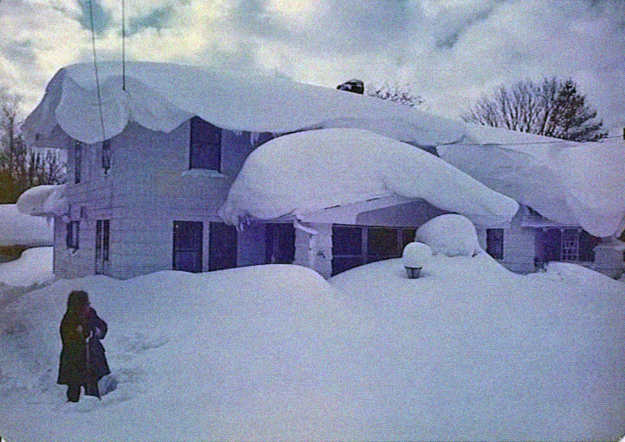 A person stands in deep snow near a house almost entirely buried under heavy snow, with large drifts covering the roof and windows. Bare trees are visible in the background under a cloudy sky.
