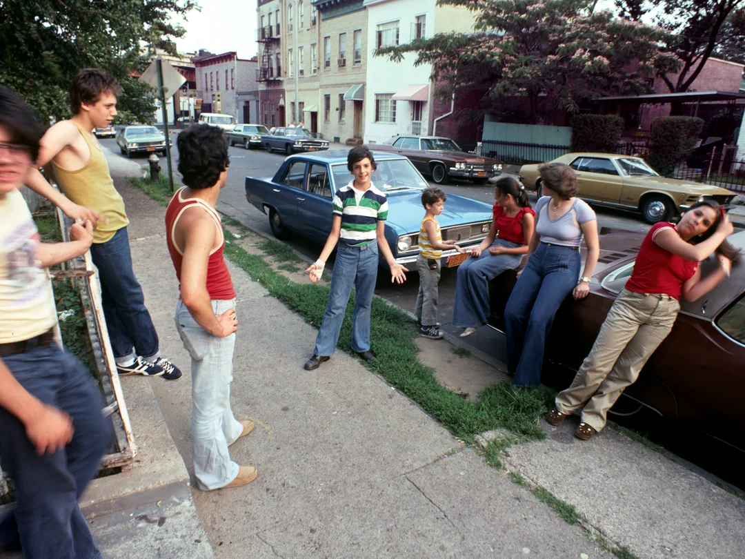 A group of teenagers in 1970s-style clothes hang out on a city sidewalk and lean against parked cars, chatting and laughing on a sunny day in a residential neighborhood.