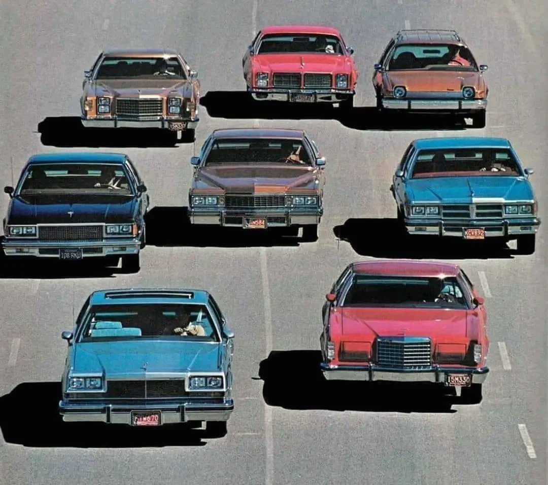 Nine classic American cars from the 1970s and 1980s are driving side by side on a wide road, arranged in a V-shaped formation. The cars are in various colors including blue, red, and tan.