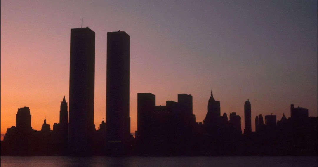 Silhouette of the New York City skyline at sunset, prominently featuring the Twin Towers of the World Trade Center against an orange and purple sky.