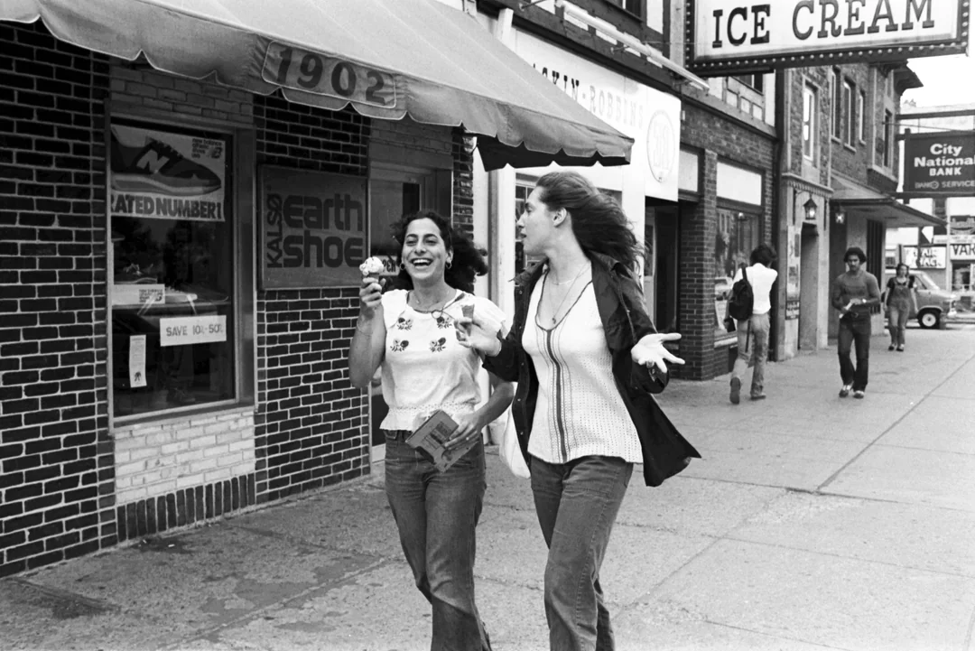 Two young women walk down a city sidewalk smiling and eating ice cream cones. Shops, including an ice cream store and a shoe store, line the street. Other pedestrians are visible in the background.