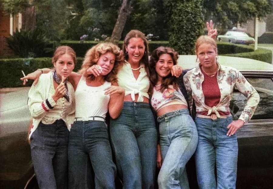 Five young women in 1970s-style clothing pose together in front of a car, smiling and making playful gestures, with greenery and houses in the background.