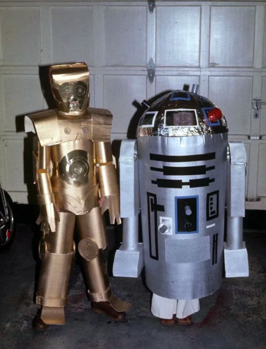 Two people in homemade costumes resembling C-3PO and R2-D2 from Star Wars stand side by side in a garage with a white paneled door behind them.