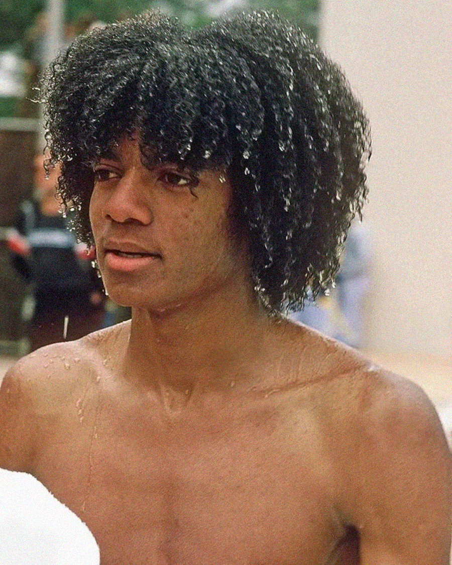 A young person with wet curly hair and droplets on their face and shoulders stands shirtless outdoors, possibly after swimming, with a blurred background.