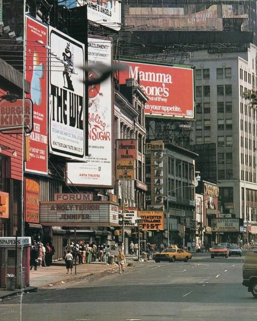 A busy city street scene with large colorful billboards, including ads for "The Wiz" and "Mamma Leone's," pedestrians on the sidewalks, and a yellow taxi driving by. Buildings line both sides of the street.