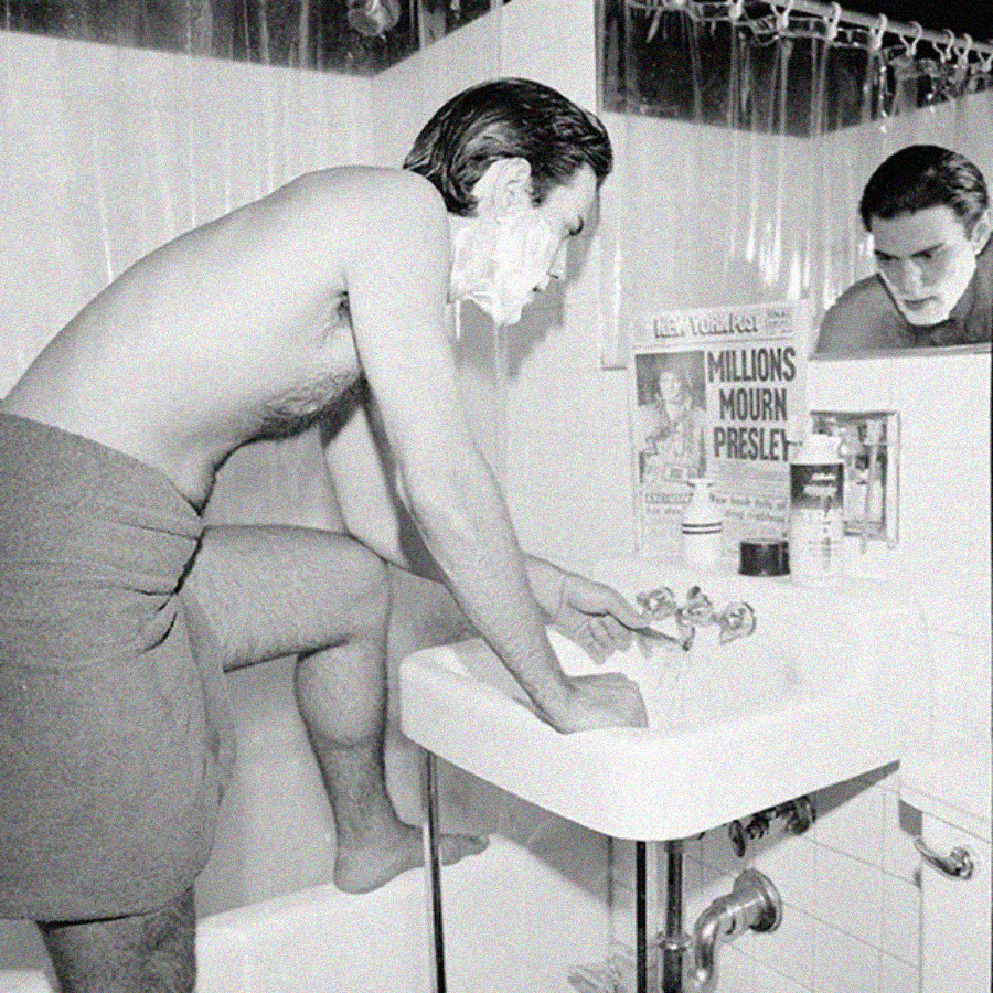 A man with shaving cream on his face stands in a bathroom, leaning over a sink with one leg on the edge. He is wrapped in a towel. A newspaper with the headline "Millions Mourn Presley" is visible by the mirror.