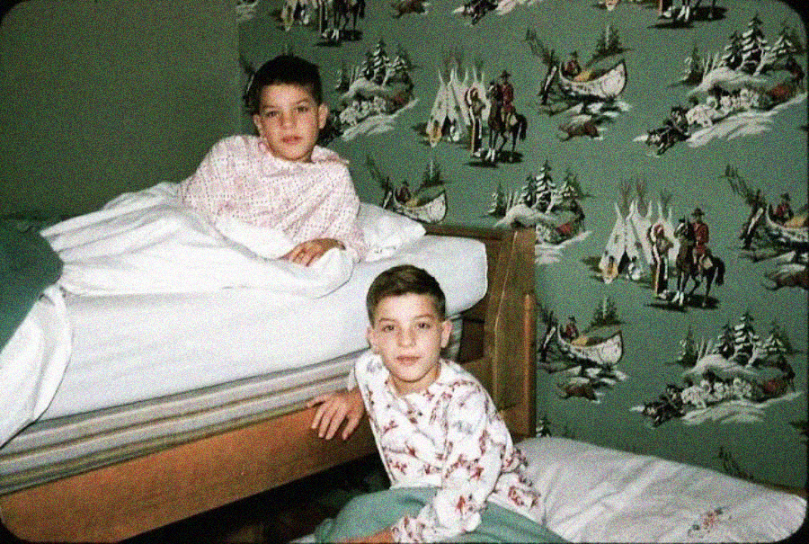 Two young boys in pajamas, one sitting on a bed and the other on a mattress on the floor, in a room with green wallpaper featuring western scenes with cowboys and horses.