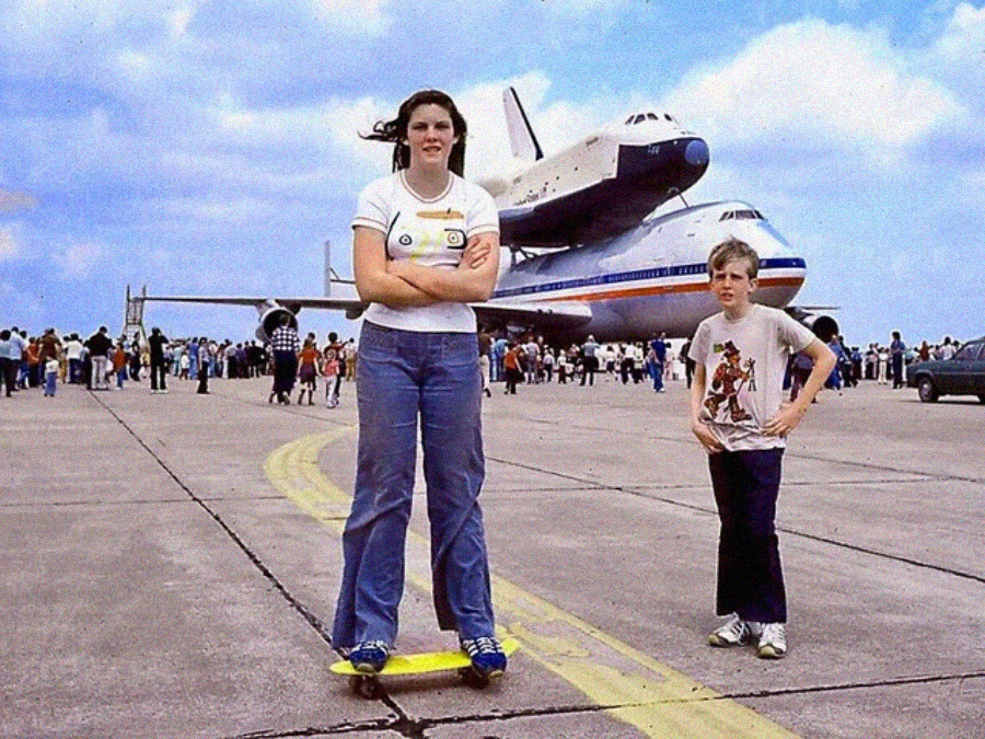 A teenage girl and a young boy pose on an airport tarmac, with a space shuttle mounted atop a NASA jumbo jet in the background. The sky is blue with scattered clouds and people are gathered around.