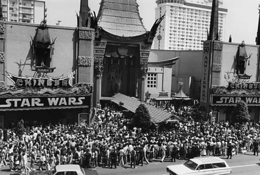 A large crowd lines up outside Mann’s Chinese Theatre for the original "Star Wars" movie premiere. The iconic theater marquee displays "STAR WARS" as people fill the sidewalk and street, with cars parked in front.