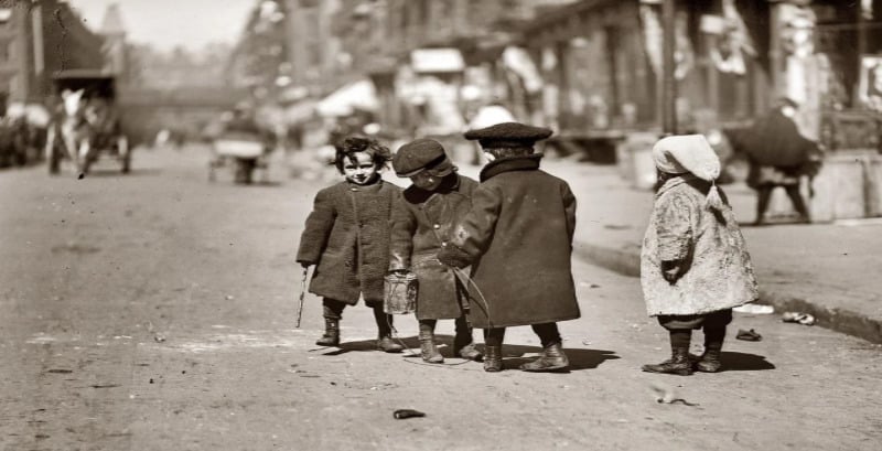 Four young children wearing coats and hats stand in the middle of a city street, facing each other. The background shows blurred buildings, sidewalks, and a horse-drawn carriage. The scene appears old-fashioned.