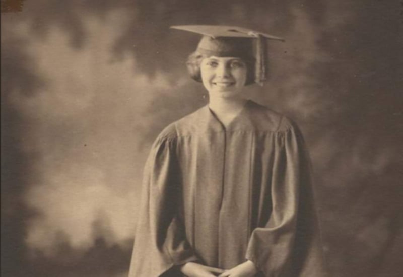 A young woman in a graduation cap and gown smiles at the camera in a sepia-toned, vintage photograph with a blurred studio backdrop.