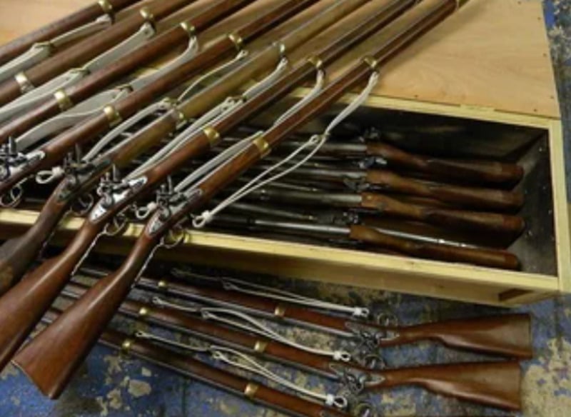 Several vintage muskets with wooden stocks are arranged on top of and inside a wooden crate, placed on a blue and brown speckled floor.