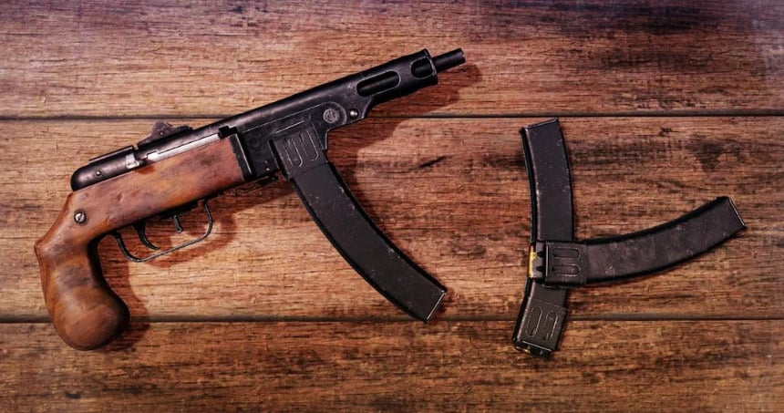 A submachine gun with a wooden grip lies on a wooden surface next to two curved, black magazines arranged in a "V" shape.