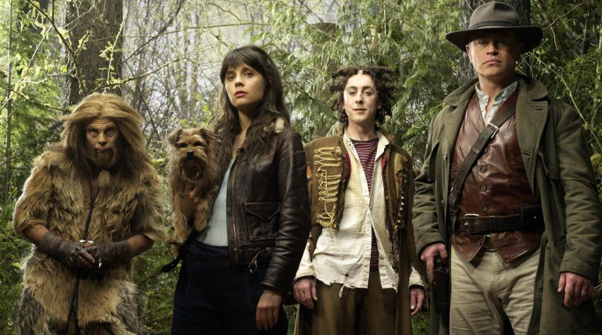 Four people in fantasy costumes stand in a forest: a person dressed as a lion, a woman holding a small dog, a person in patchwork clothes, and a man in a leather coat and wide-brim hat, all looking ahead.
