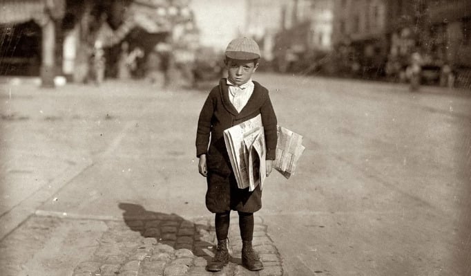 A young boy wearing a cap and dark clothes stands on a street holding several newspapers under his arm. The background is a blurred city scene with buildings and people.