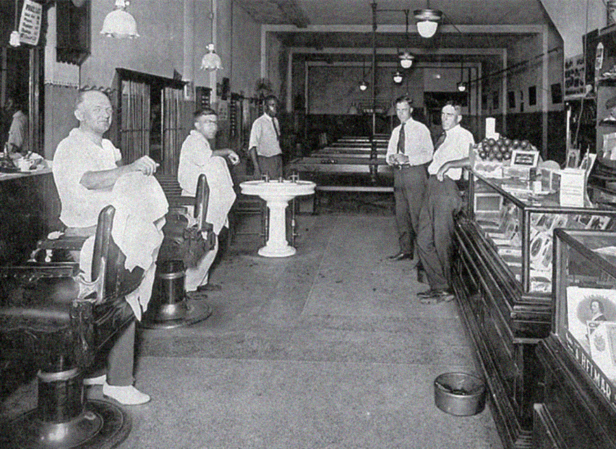 Black and white photo of a vintage barbershop with five men inside. Three are barbers in white coats near barber chairs, and two men stand near a counter with display cases. The shop has high ceilings and hanging lights.