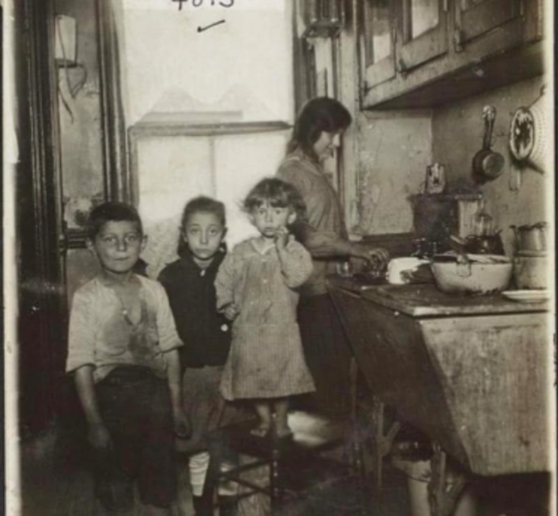 Three children stand in a small, cluttered kitchen while a woman, likely their mother, works at the sink. The room appears old and worn, with various kitchen items on the counter and hanging on the wall.