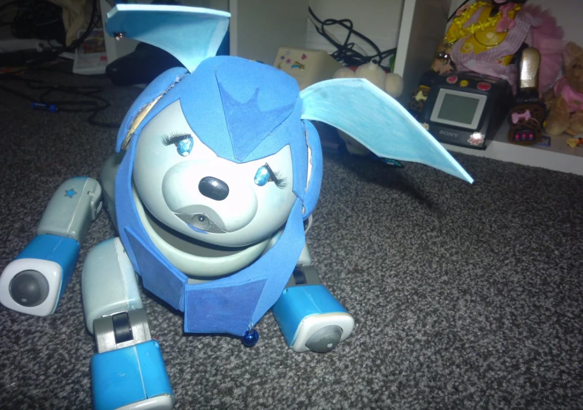 A robotic dog toy with blue ears, blue limbs, and a blue headpiece sits on a gray carpet. Shelves with toys, cords, and electronic devices are visible in the background.
