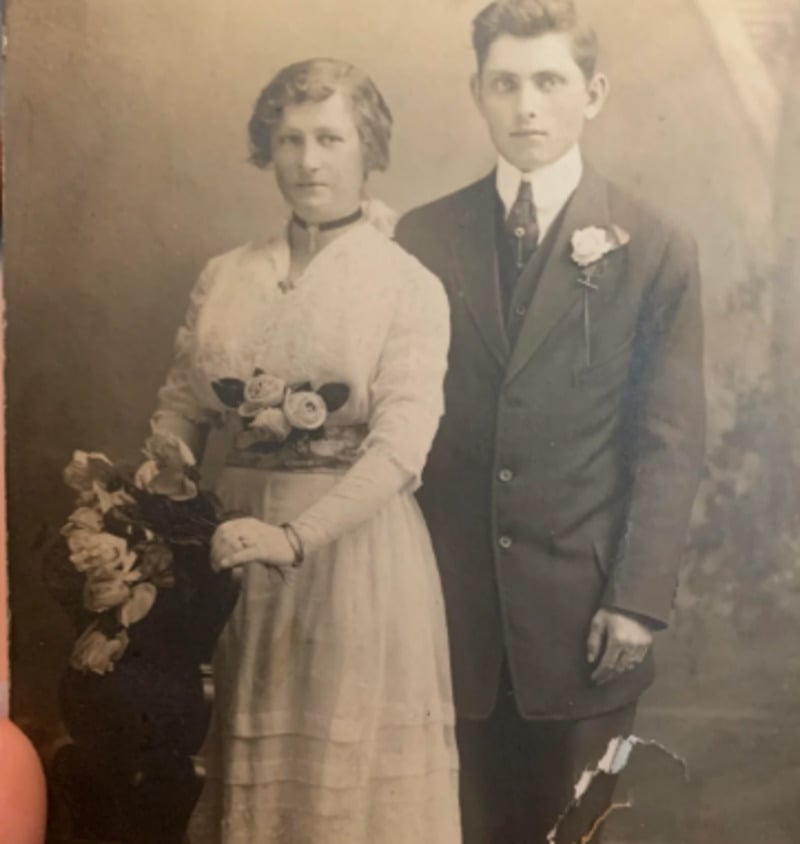 A sepia-toned vintage photograph of a woman in a long white dress with floral details and a man in a dark suit with a boutonnière, standing side by side, likely from the early 20th century.
