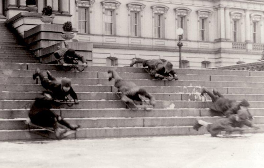 A black-and-white photo shows several people riding sleds down wide stone steps outside a grand building, appearing mid-motion and somewhat chaotic. The scene is lively and historic.