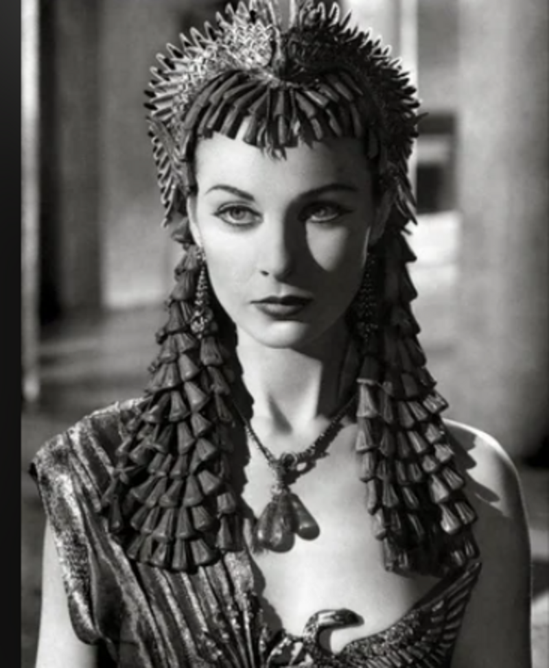 A woman in elaborate ancient Egyptian-inspired attire, wearing a decorative headdress, statement earrings, and a patterned dress, gazes forward in this black-and-white photograph.