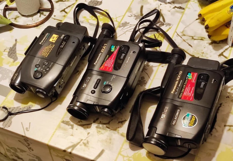 Three vintage Panasonic camcorders are placed side by side on a patterned tablecloth. A bunch of bananas and a metal lid are visible in the background.