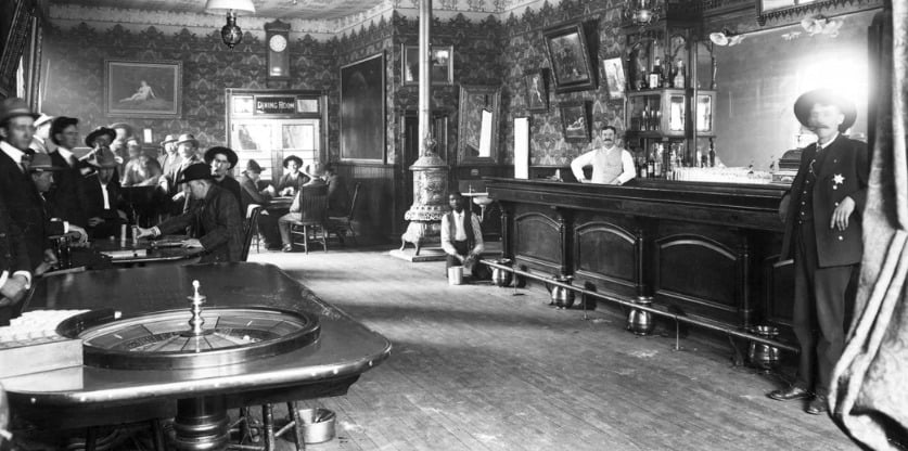 Black-and-white photo of an old Western saloon with men in hats, a bartender behind a long bar, a roulette table in front, and ornate wallpaper and furnishings throughout the room.