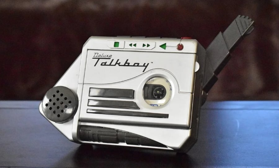 A silver Deluxe Talkboy cassette recorder with a built-in microphone, playback buttons, and an extended microphone arm, sitting on a dark surface.