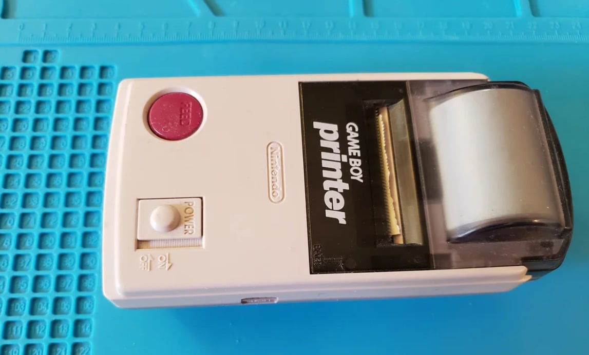 A white Nintendo Game Boy Printer with a red feed button and clear paper cover, placed on a blue surface.