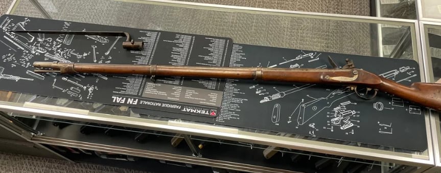 A long antique rifle with a wooden stock and metal barrel is displayed on a glass counter, alongside a detached ramrod and cleaning rod, atop a gun schematic mat.