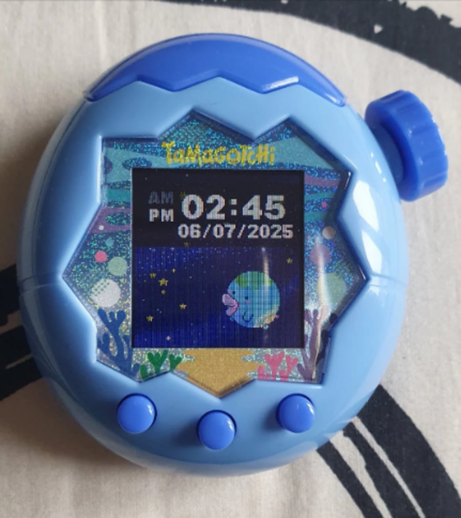A blue Tamagotchi device displays the time 02:45 PM and the date 06/07/2025. The screen shows an animated fish character with underwater scenery and star patterns in the background.