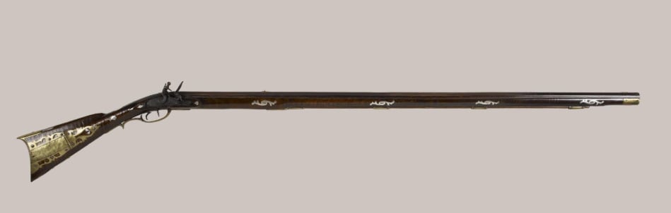A long, antique flintlock rifle with a dark wooden stock and brass inlays, featuring decorative metalwork along the stock and barrel, displayed on a plain light background.