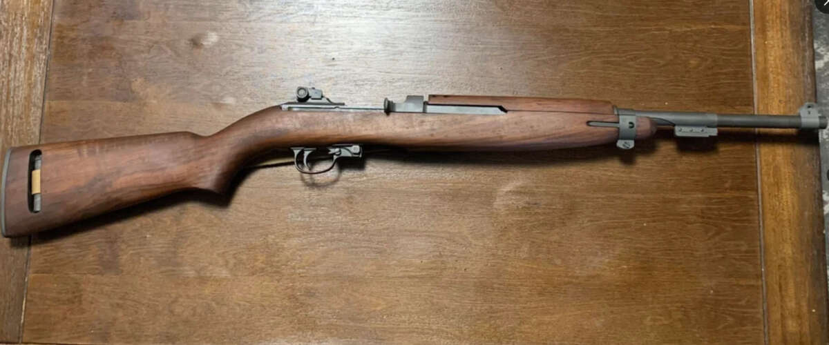 A vintage M1 Carbine rifle with a wooden stock is displayed on a wooden surface, showing the entire firearm from butt to barrel.