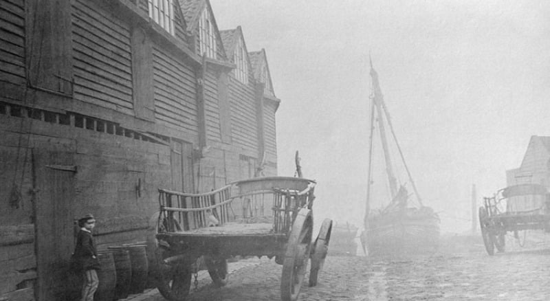 A black-and-white photo shows a cobbled street with wooden warehouses on the left, an empty horse-drawn cart, a person standing nearby, and a docked sailing ship in the foggy background.