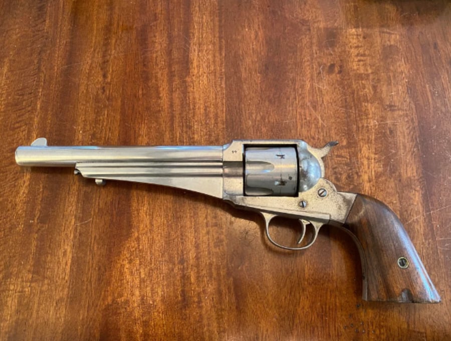 A silver revolver with a long barrel and a wooden grip rests on a wooden surface. The revolver is displayed flat, showing its side profile.
