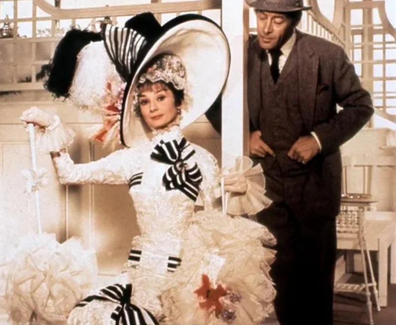 A woman in an elaborate white dress with black stripes, matching hat, and parasol poses elegantly as a man in a suit and bowler hat stands behind her, both in a richly decorated, vintage setting.