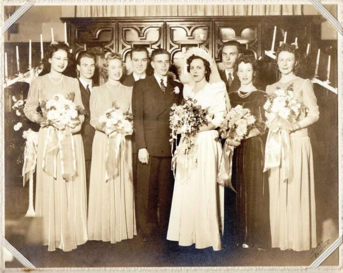 A vintage sepia photo of a wedding party: bride and groom in the center, flanked by bridesmaids holding bouquets with ribbons and groomsmen in suits, standing in front of a wooden backdrop with lit candles.