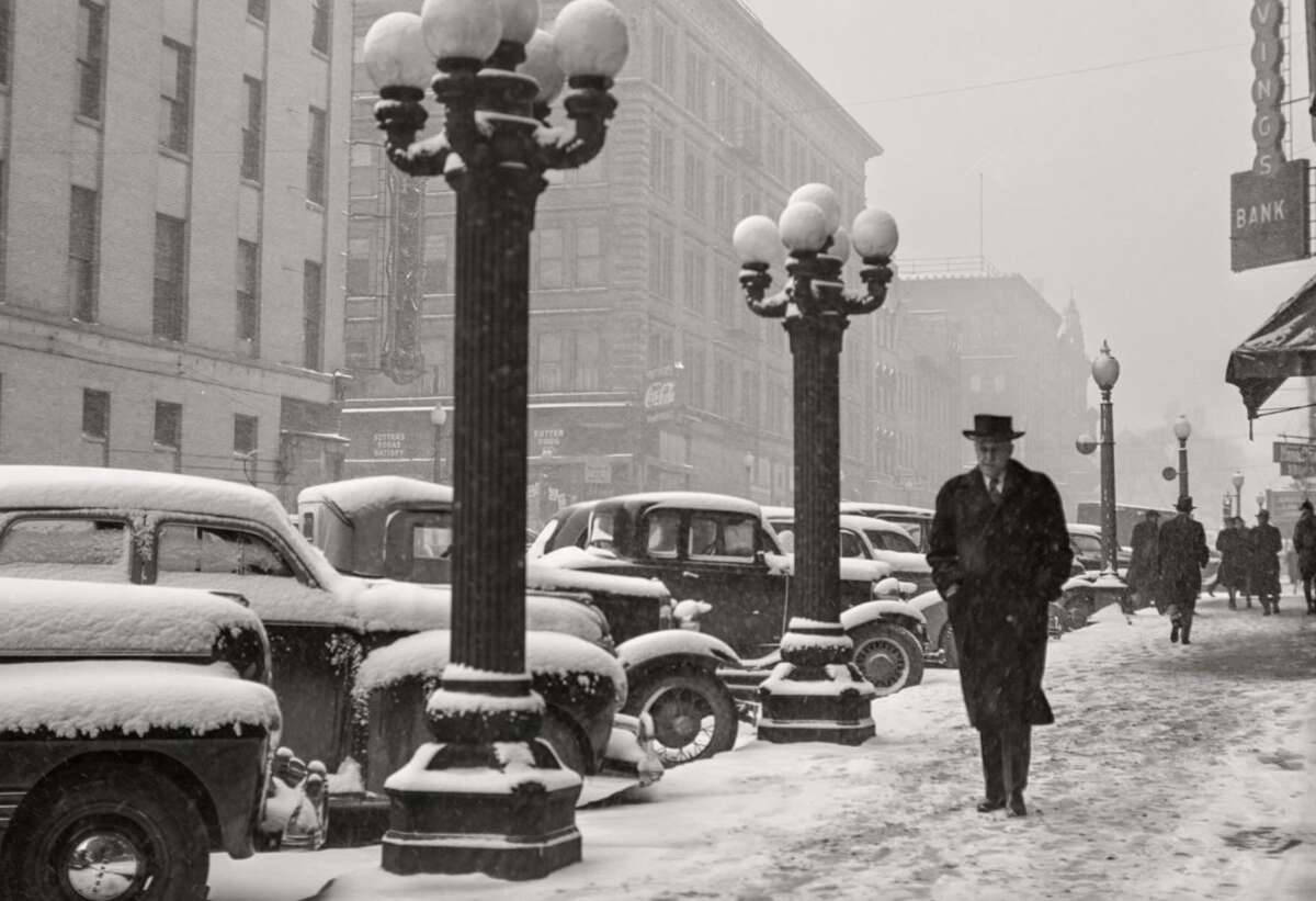 Black-and-white photo of a snowy city street lined with parked vintage cars and ornate street lamps. People in coats and hats walk along a snow-covered sidewalk. Buildings and a bank sign are visible in the background.