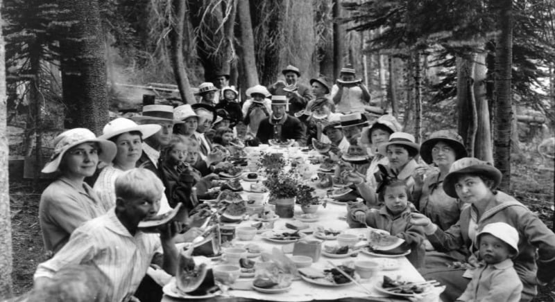 A black-and-white photo of men, women, and children in hats sitting at a long outdoor table in a forest, sharing a meal together. The table is covered with plates of food and drinks.